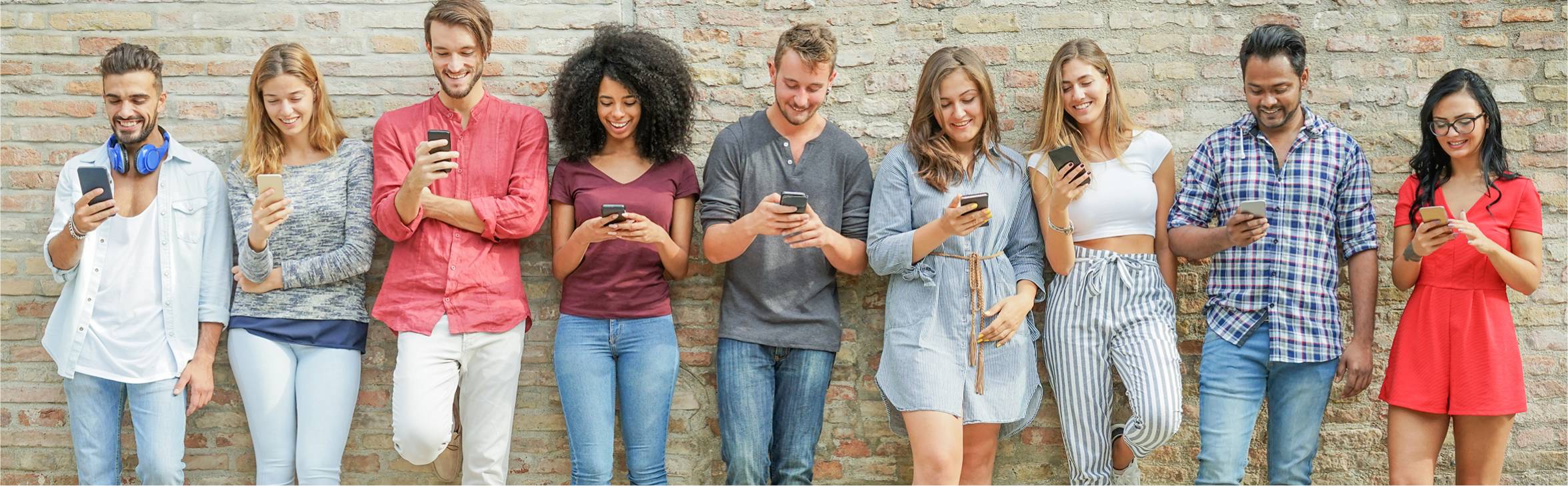 A group of people looking at their phones while standing against a wall.