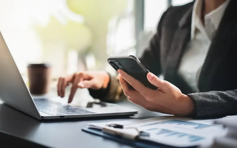 Woman holding phone and using laptop