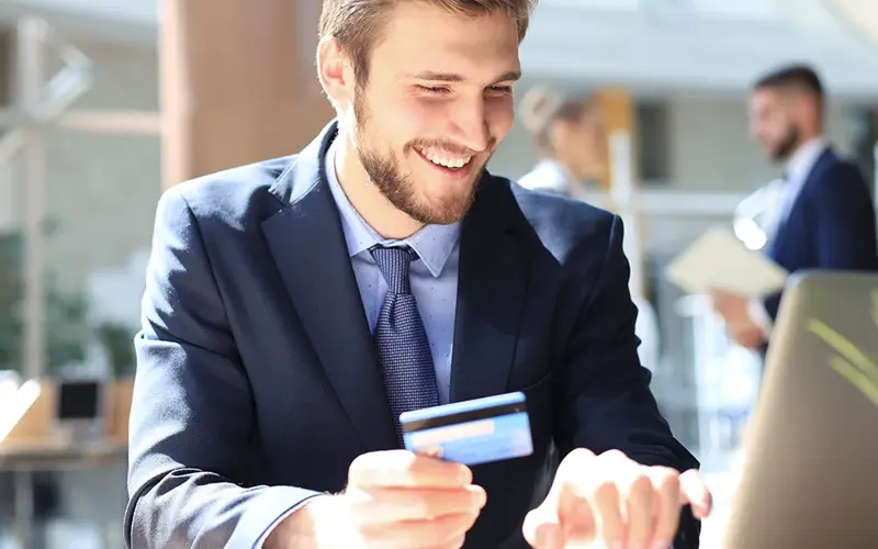 Smiling man sitting in office and pays by credit card with his laptop