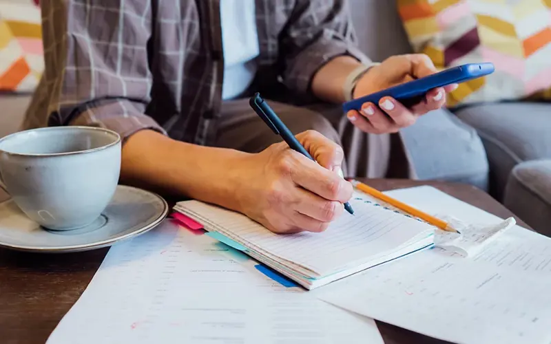 Woman reviewing expenses, managing personal finances on smartphone mobile app while sitting on couch with coffee