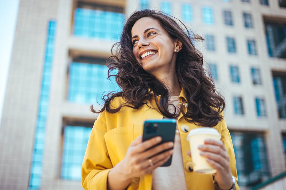 Young positive businesswoman with take away coffee cup in hand using mobile phone while standing on sunny street