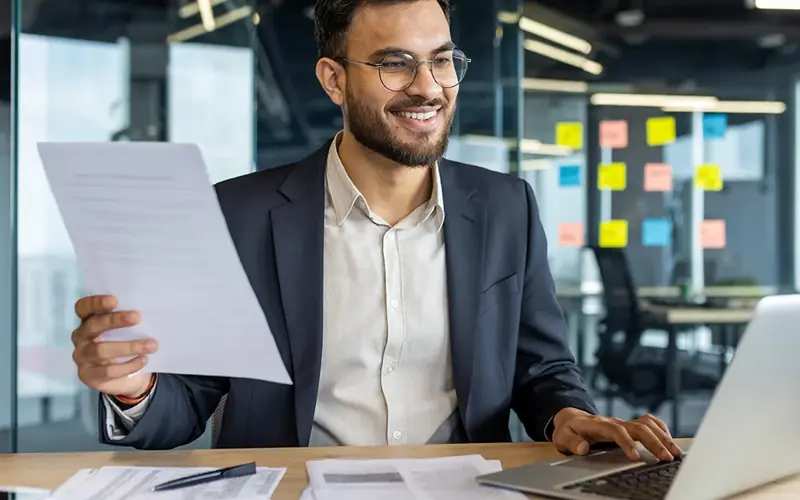 A smiling businessman reviews documents while working on a laptop