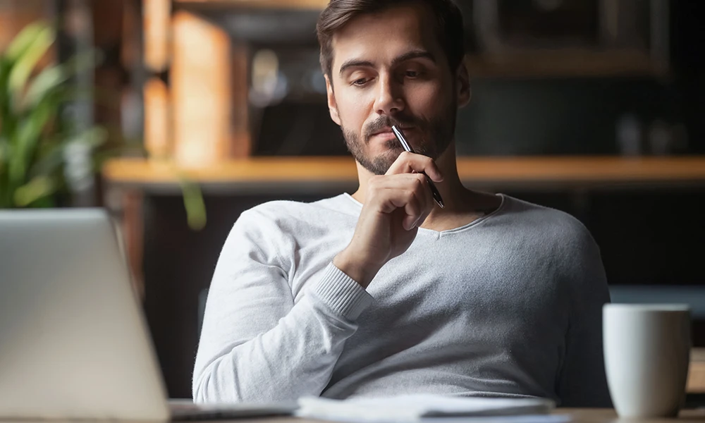 man holding pen while pondering what he sees on his computer