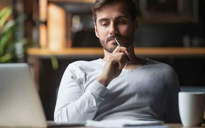 man holding pen while pondering what he sees on his computer