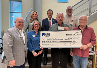A group of F&M Trust and Fulton County Medical Center employees with Richard Barnett smiling and holding a large presentation check and a normal check showing both portions of the donation.