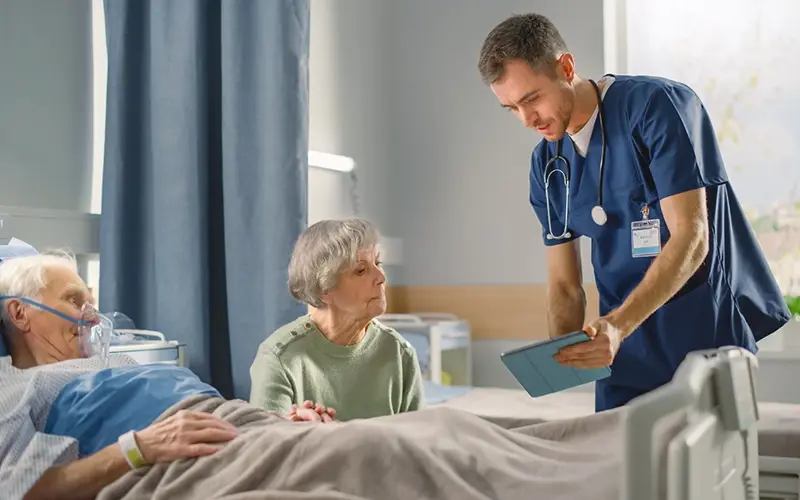 older woman looking at paperwork while incapacitated husband is in hospital bed