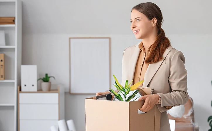 a business woman smiling as she carries a box of things into her new office
