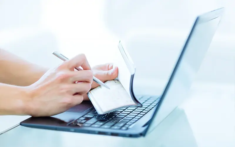 Close-up of a person's hands writing a bank cheque as a money transaction while online on a laptop computer indoors